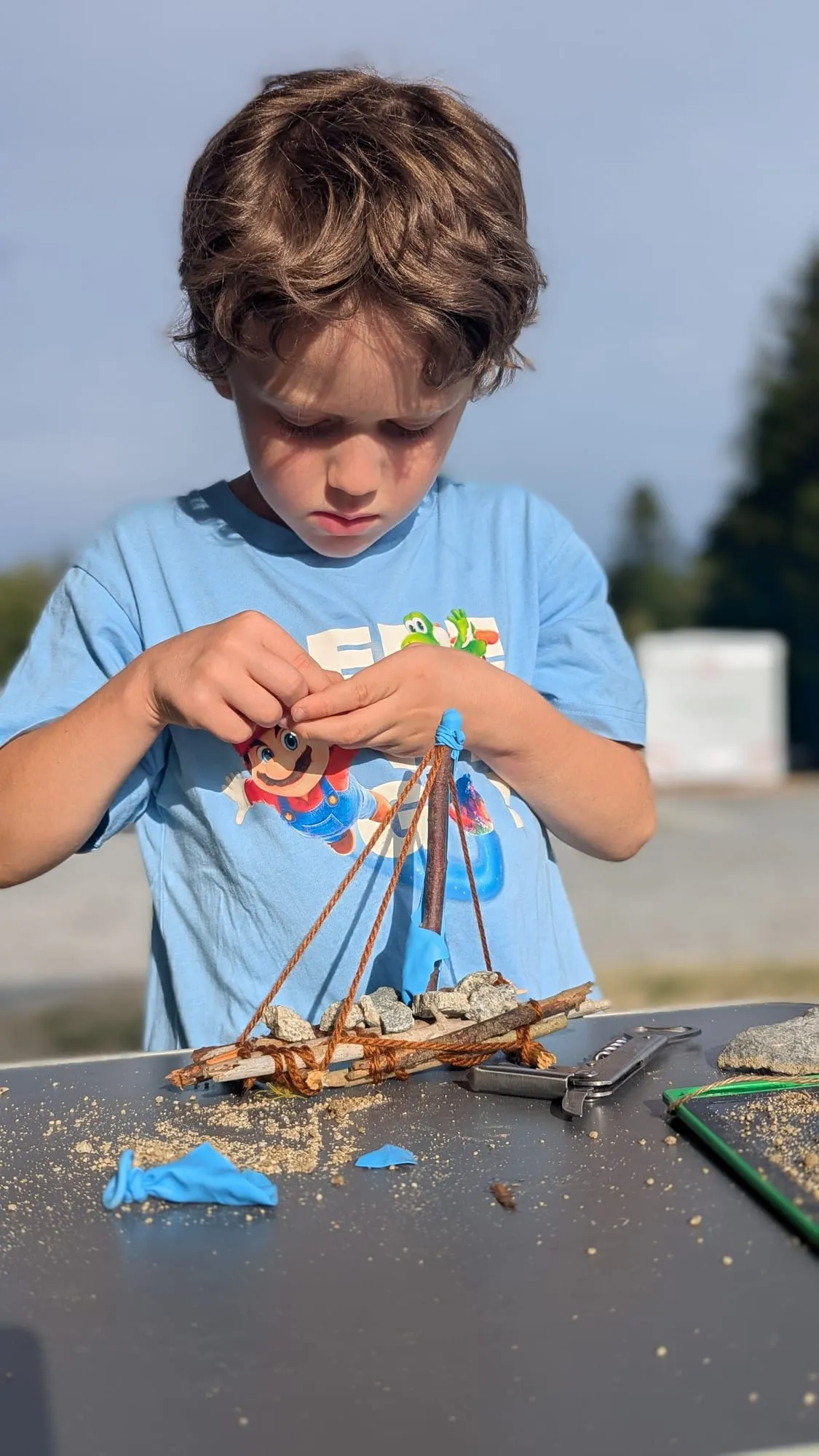Mio bindet am Campingtisch Äste mit Schnur zu einem kleinen Floß zusammen