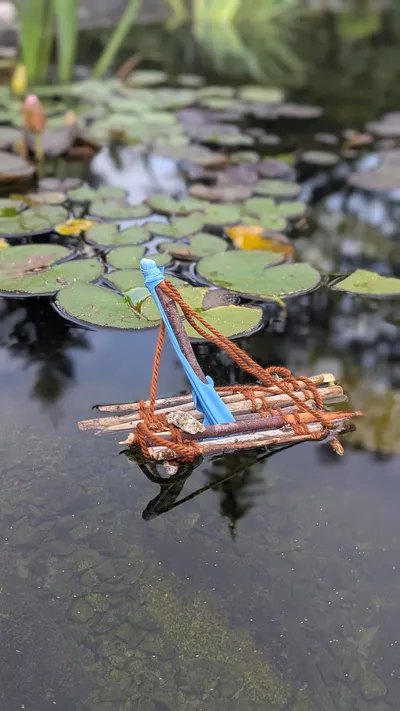 Ein selbstgebautes Mini-Floß aus Ästen schwimmt auf einem Gartenteich zwischen Seerosenblättern